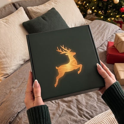 Caja de regalo verde con la silueta de un reno sobre la cama junto al arbol de Navidad